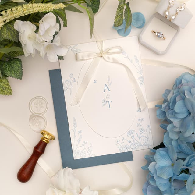 Blue floral oval wedding invitation with A & T initials, tied with an ivory satin bow on a dusty blue envelope, beside ivory wax seals, a wax seal stamp and blue hydrangeas