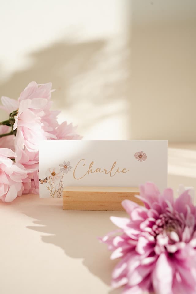 A white place card with a floral design and the name "Charlie" written in gold script. The place card is sitting on a wooden stand and is surrounded by pink flowers.