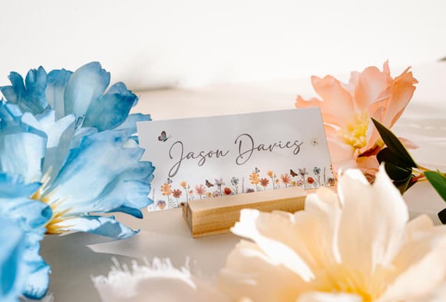 Wildflower wedding place card with the name Jason Davies in grey script above a border of watercolour meadow flowers and butterflies, standing in a wooden holder between blue, peach and cream flowers